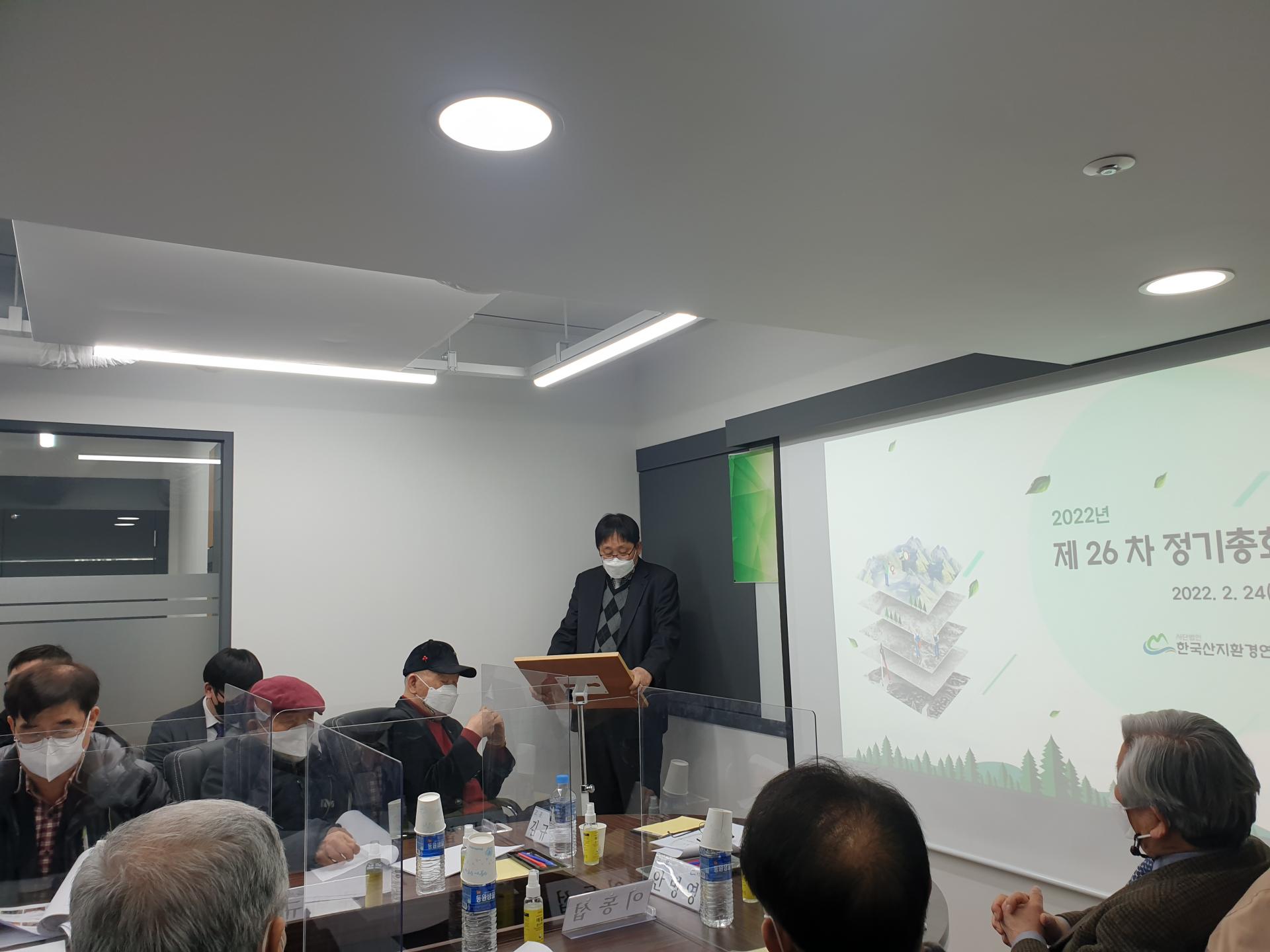 제26차 정기총회: 발표자가 2022년 총회 화면 앞에서 경과를 보고하는 모습